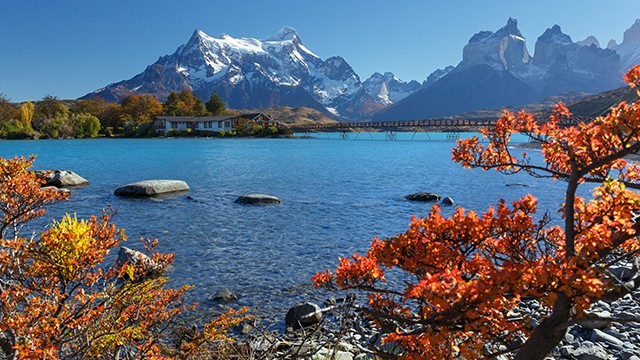 Crystal clear lake with autumn foliage, snowy mountains, and a bridge in the background.
