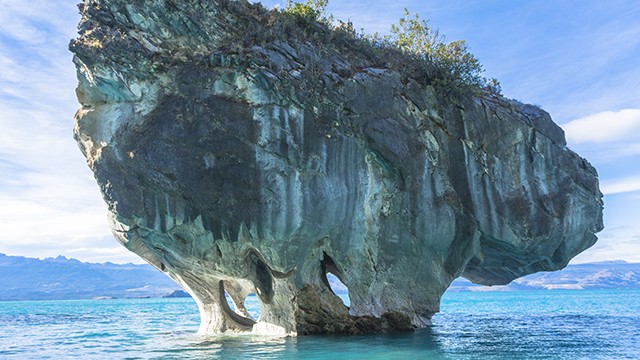 Massive, solitary marble rock formation with natural arches standing in clear blue waters.