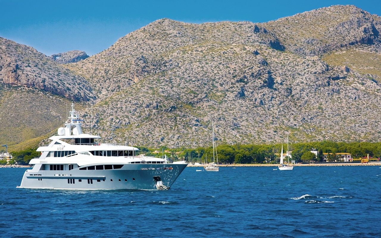 A luxury yacht sails on blue waters with a mountainous coastline in the background.