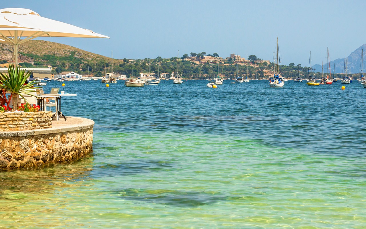 A coastal restaurant overlooks a bay with sailboats, clear waters, and a distant hillside.