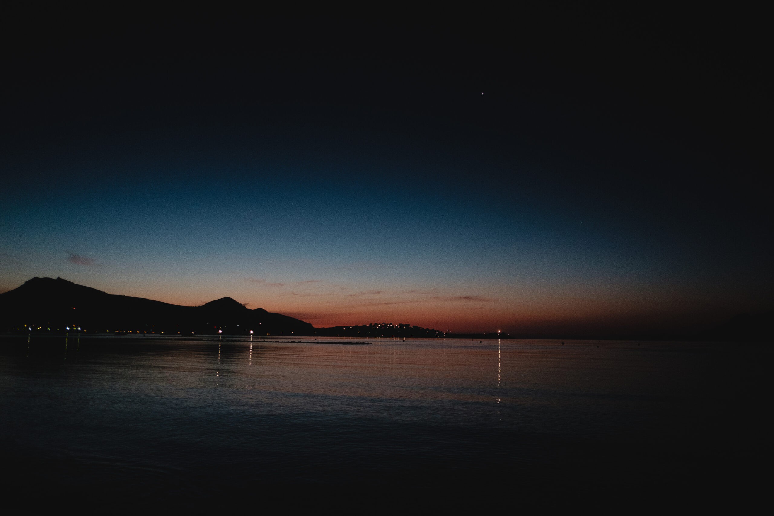 A serene twilight scene with silhouetted mountains, calm waters, and faint lights reflecting.
