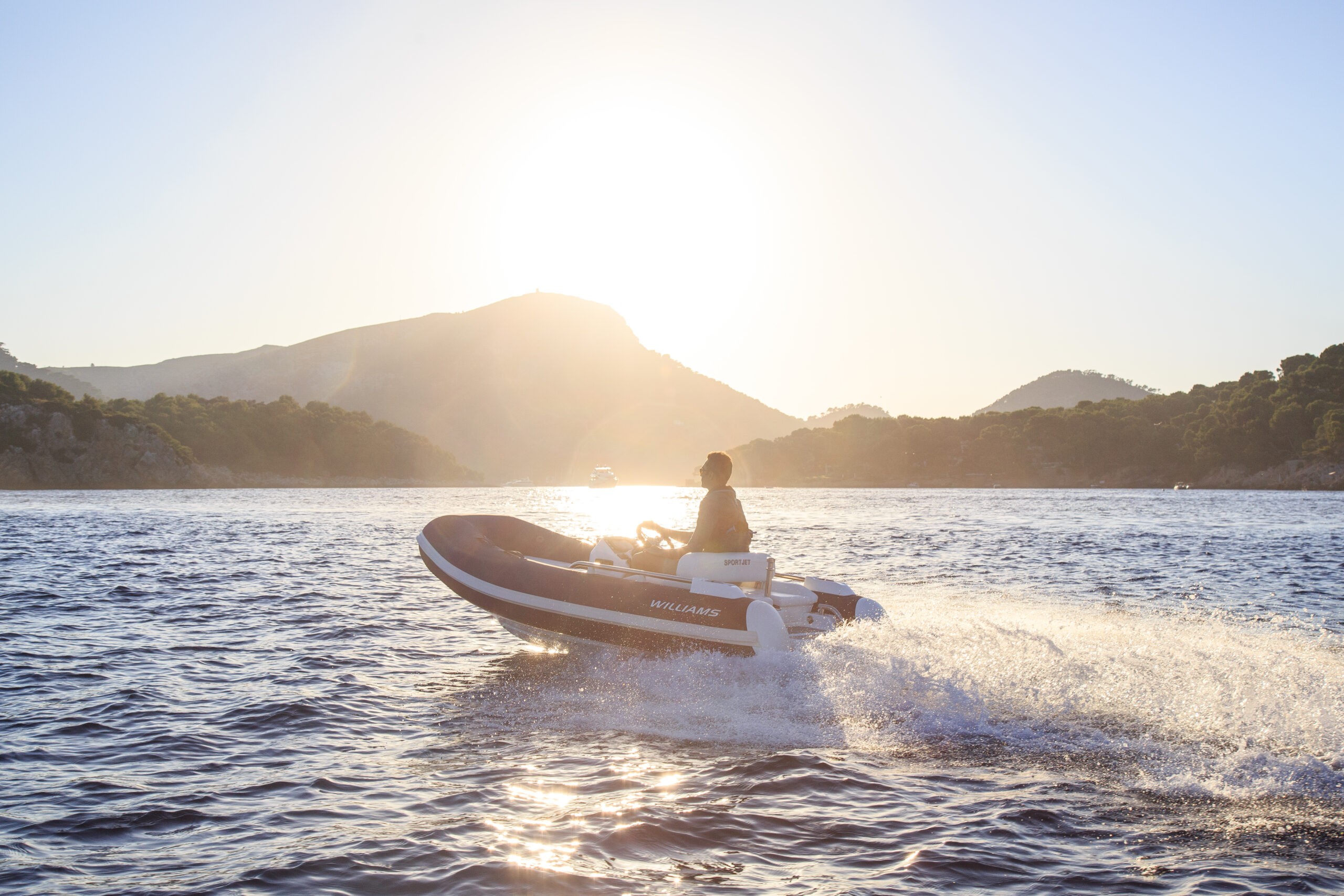 A man drives a Williams SportJet tender on a serene sea at sunset.