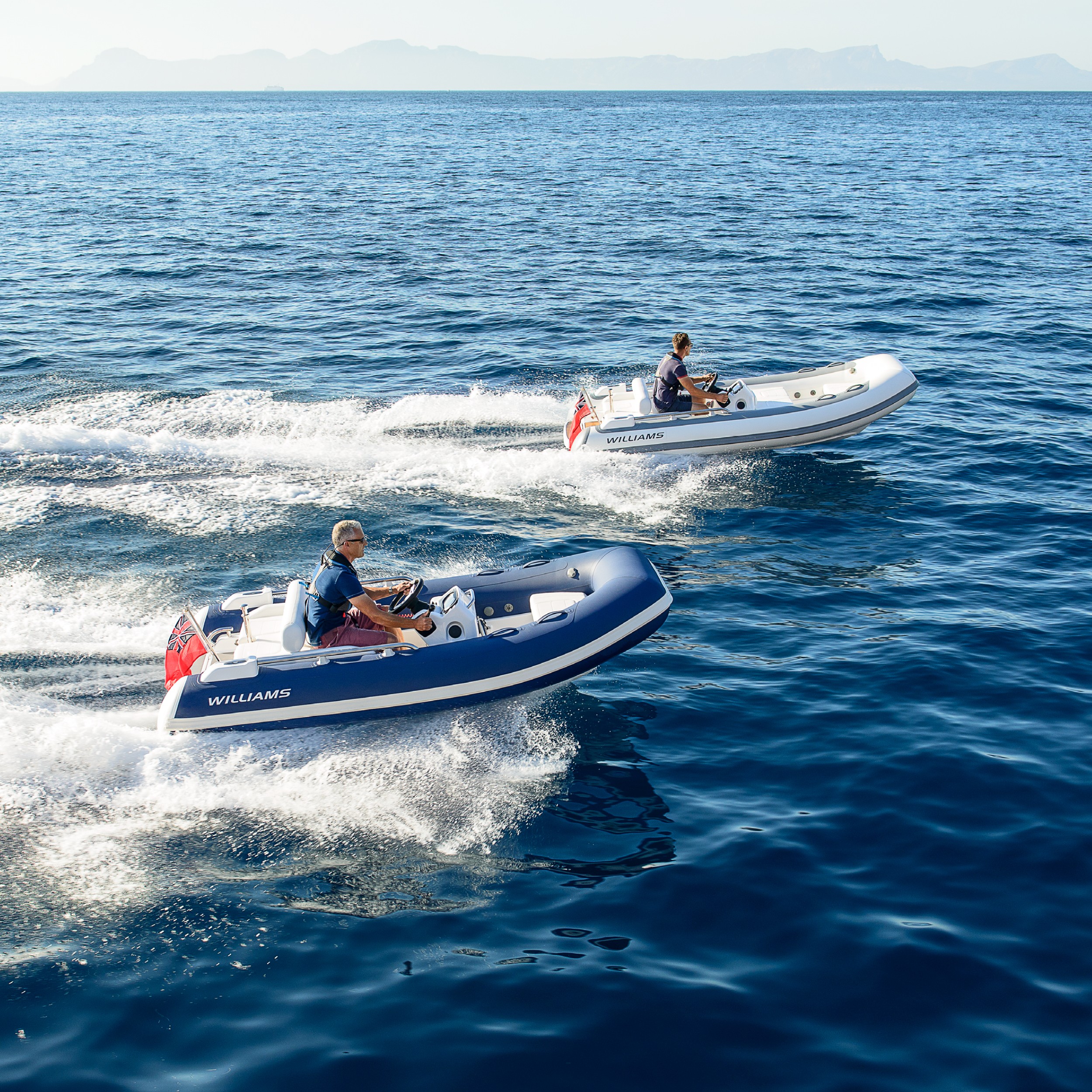 Two people drive small jet tenders on the open water with distant mountains visible.