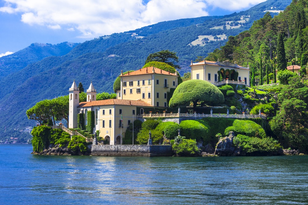 A picturesque villa with lush gardens, nestled by the lake against a mountainous backdrop.