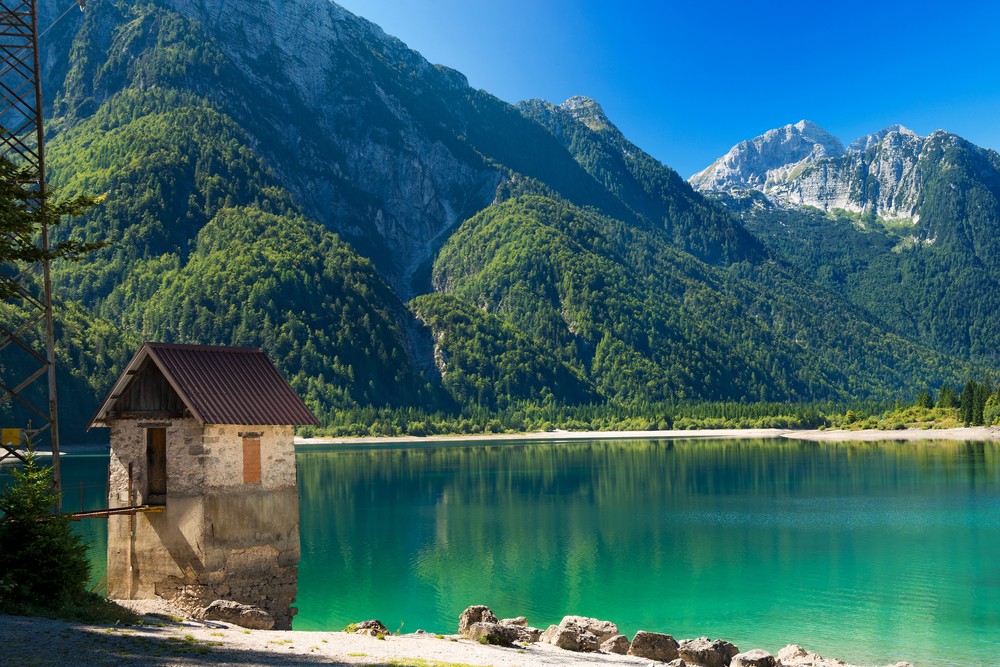 A small cabin by a tranquil, emerald lake, surrounded by lush green mountains.