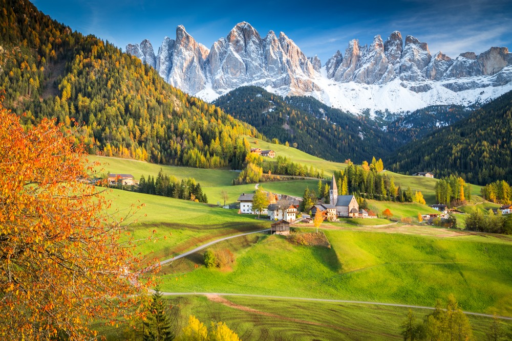 A picturesque village nestled in lush green hills with snow-capped mountains in the background.