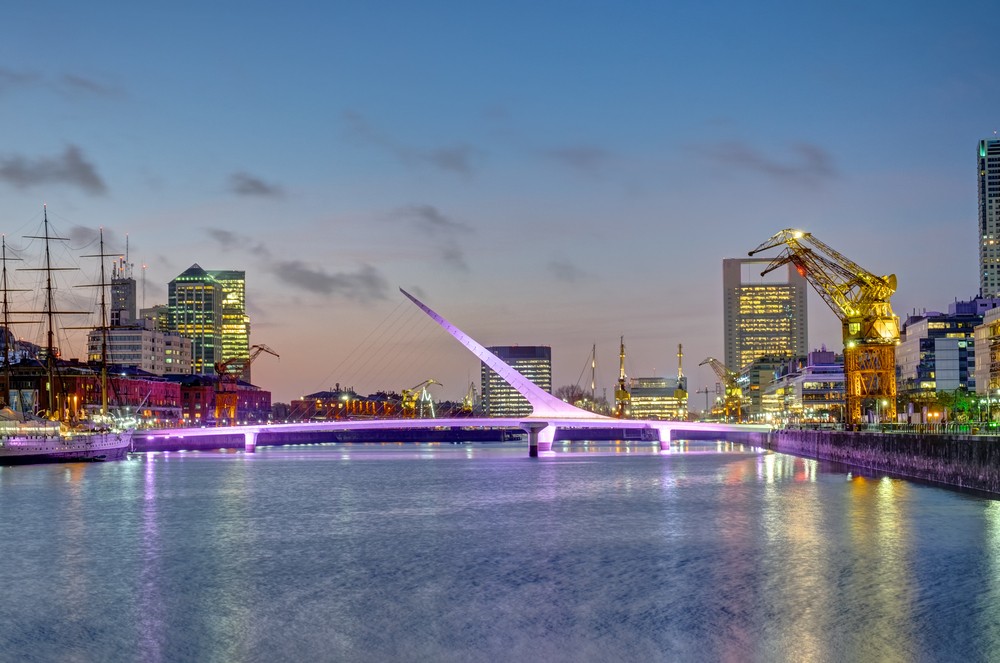 A cityscape at dusk featuring a modern bridge, illuminated buildings, and a docked ship.