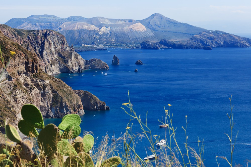 Cliffs overlooking a deep blue sea with boats and distant mountainous terrain under a clear sky.