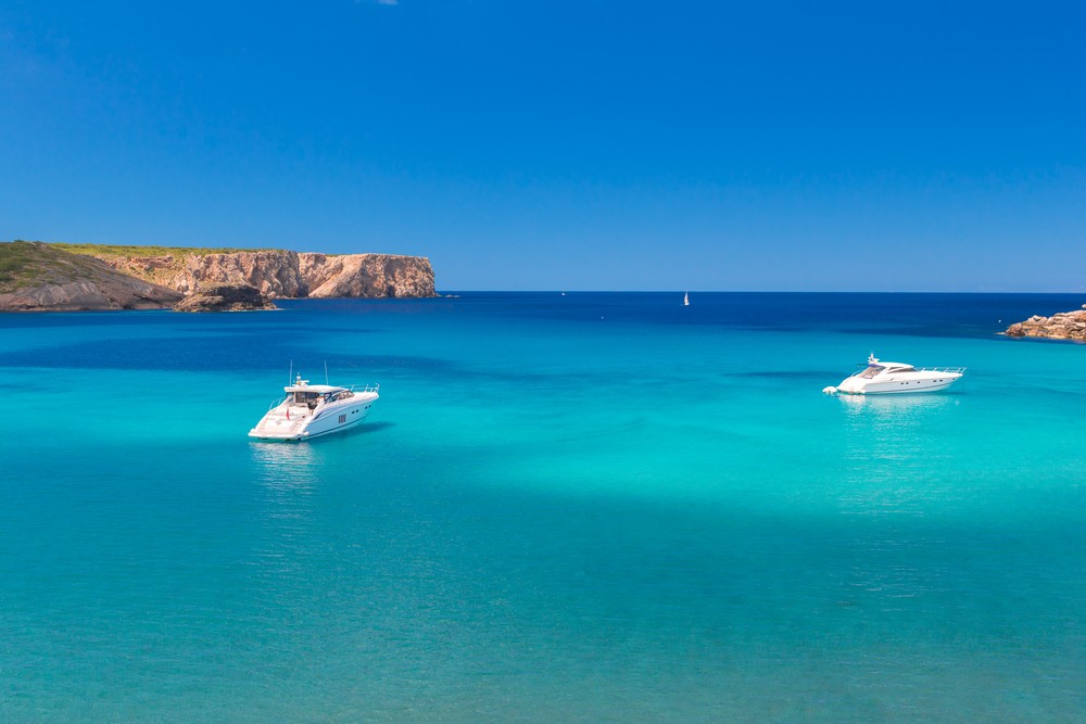 Two yachts floating on clear turquoise water with rocky cliffs in the background under a bright blue sky.