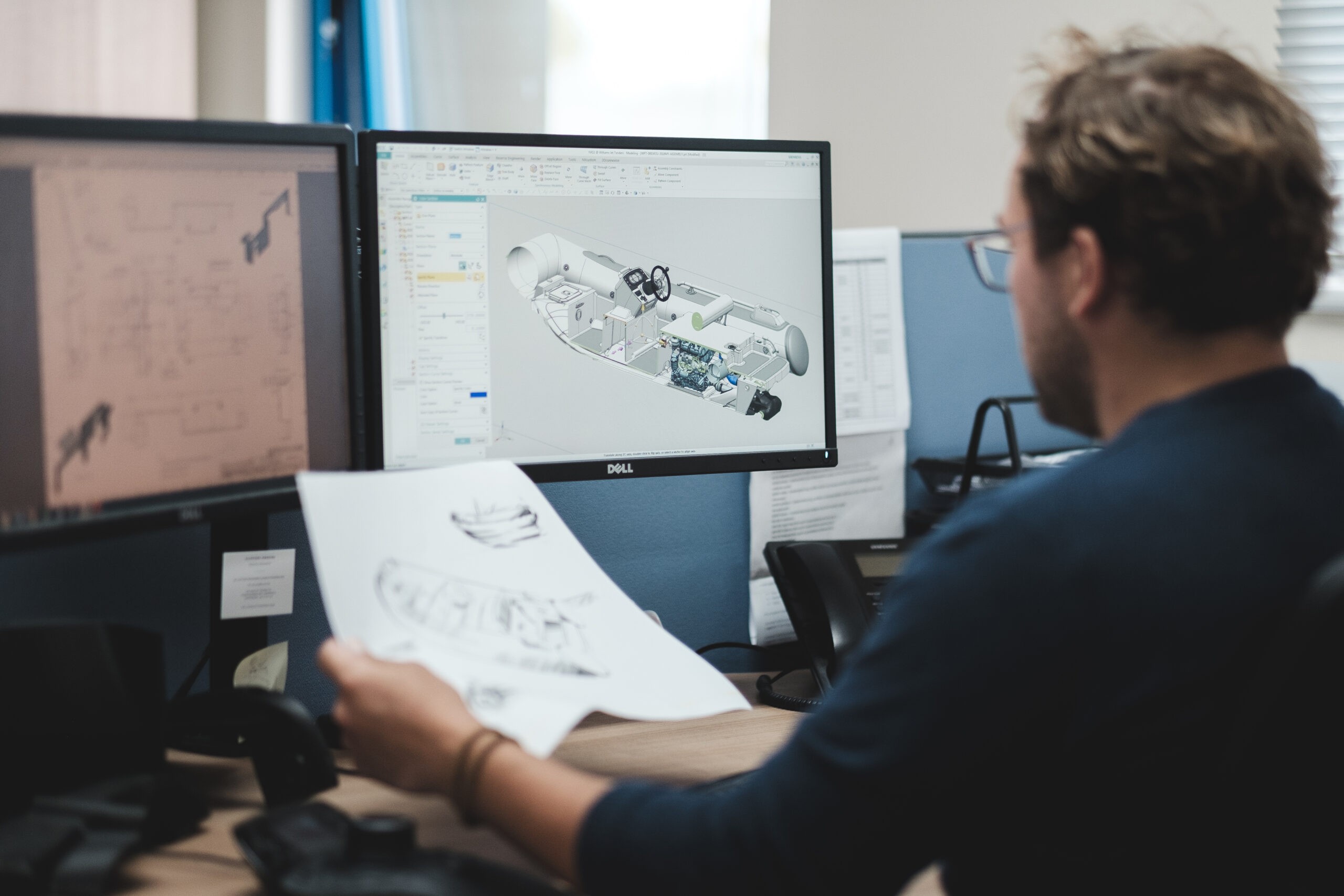 A designer working on a boat's 3D model, holding a technical drawing, in an office.