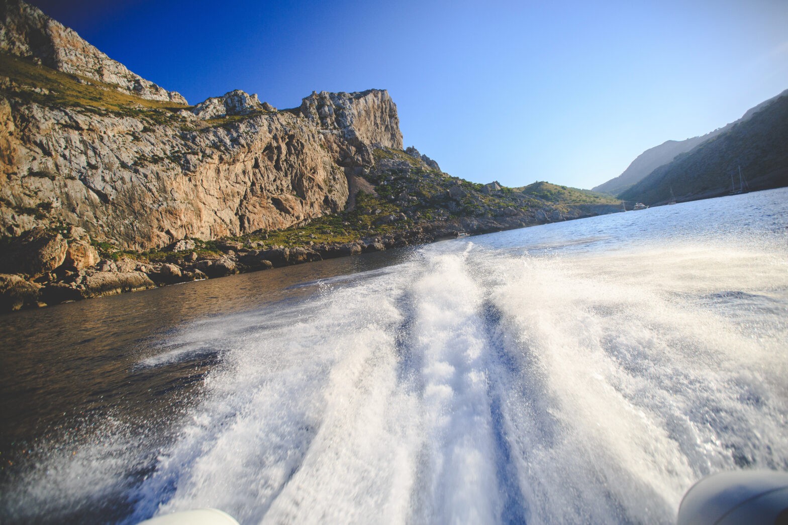A boat leaves a trail of water while cruising past rocky cliffs under a clear blue sky