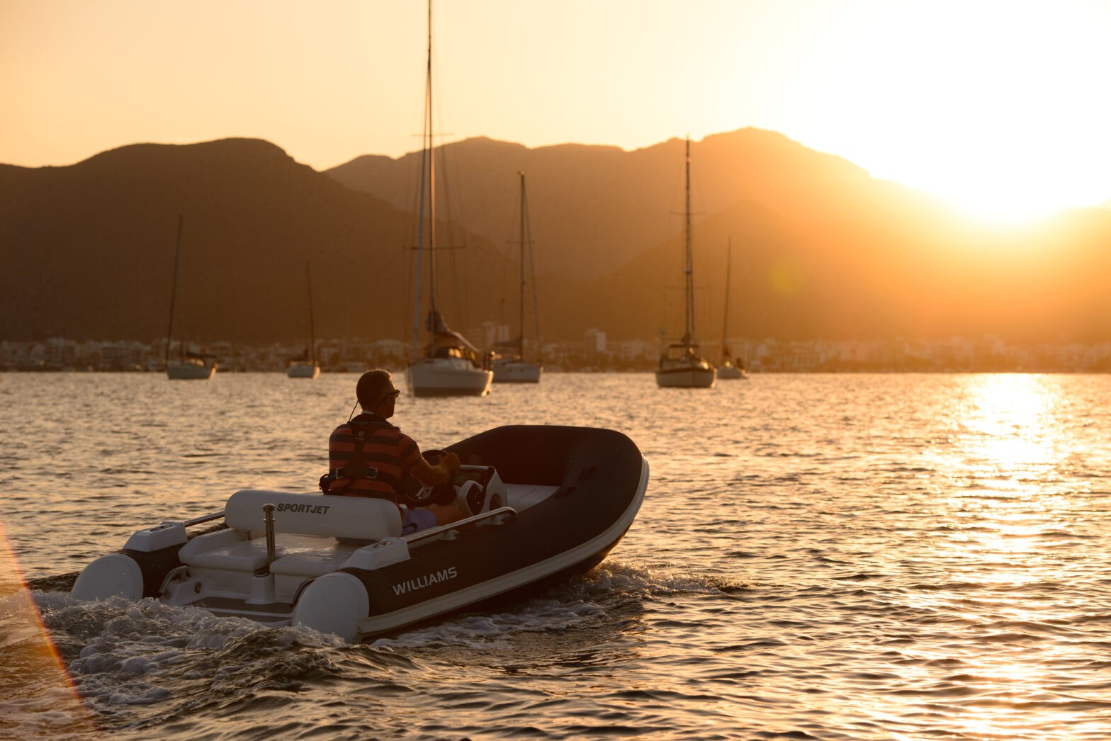 A person on a Williams yacht tender at sunset, with mountains and sailboats in the background.