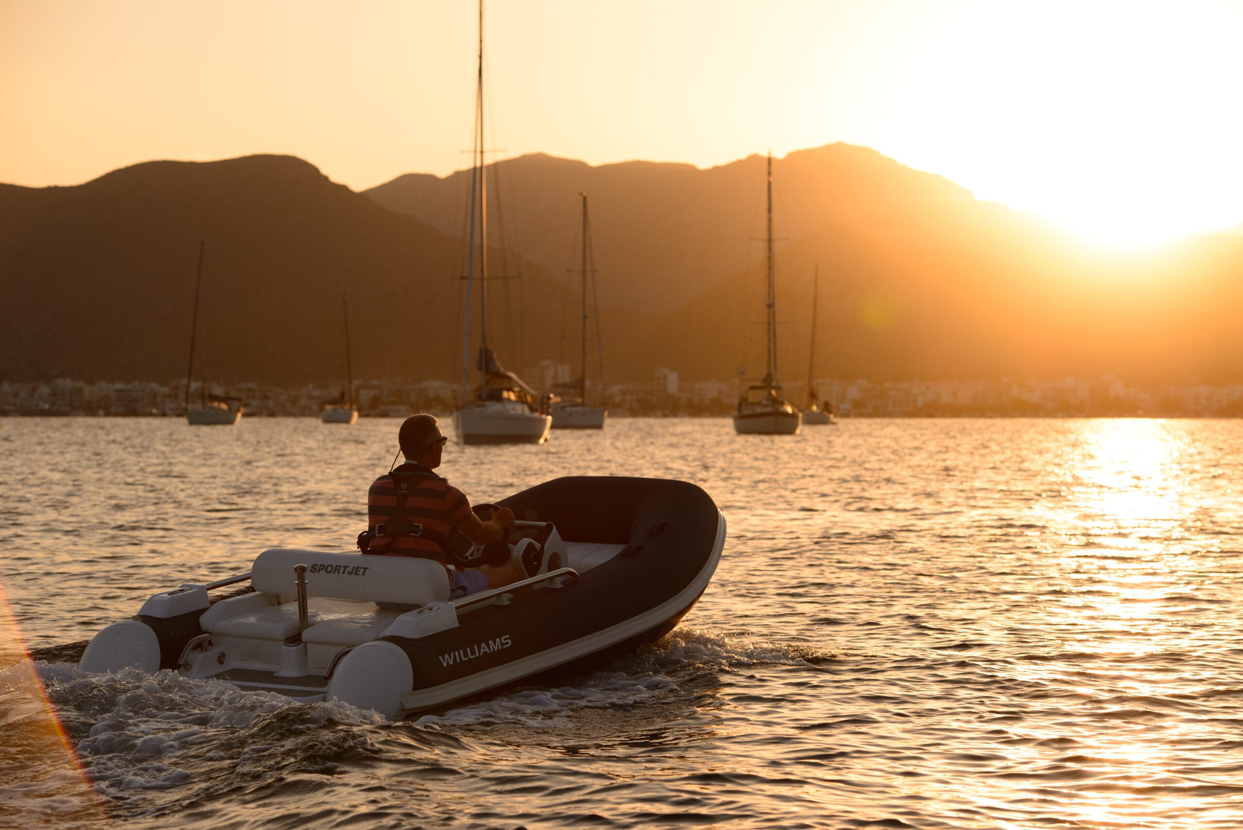 A person on a Williams yacht tender at sunset, with mountains and sailboats in the background.