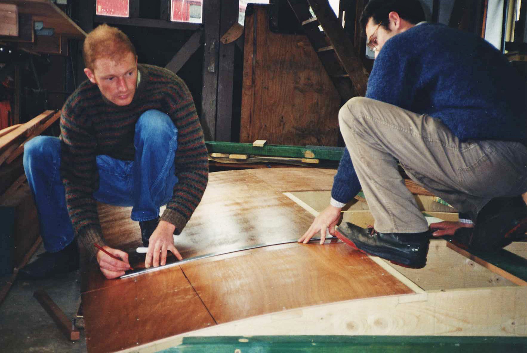 Two men measuring and marking a wooden board inside a workshop, engaged in constructing the boat.