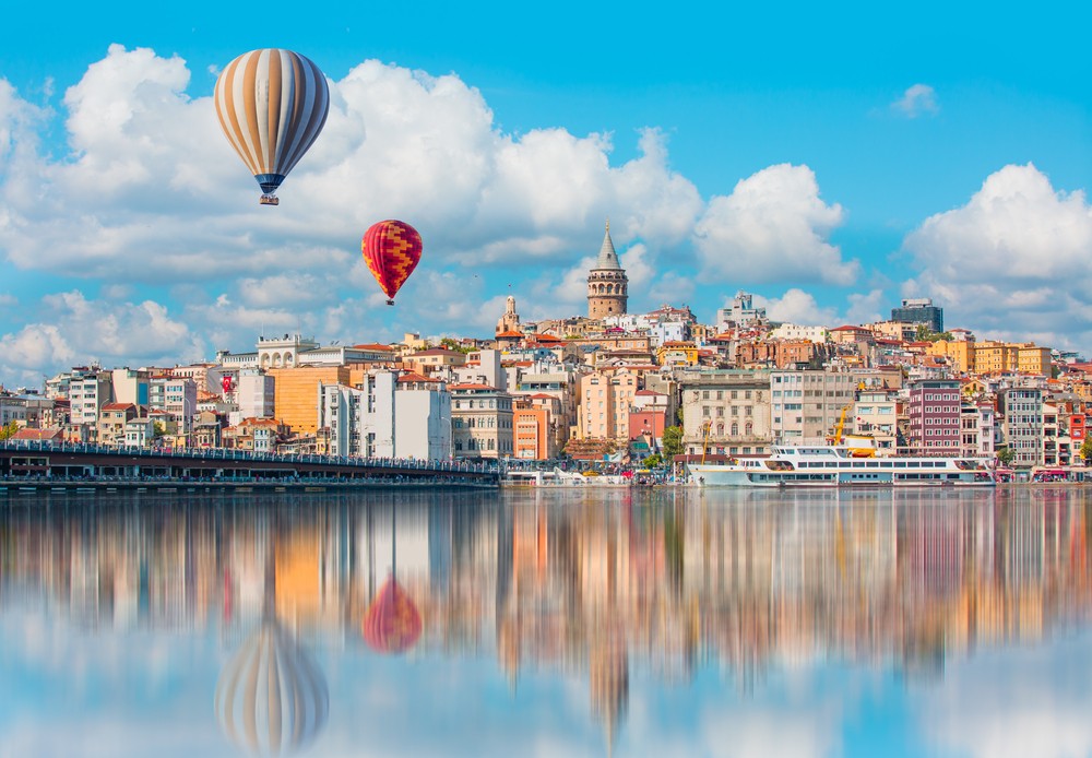 Hot air balloons float over a scenic cityscape with colourful buildings and a waterfront